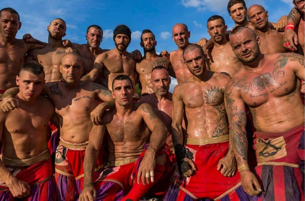 A large group of muscular adult men, many with tattoos, are standing outdoors wearing red and purple shorts. The group is smiling.
