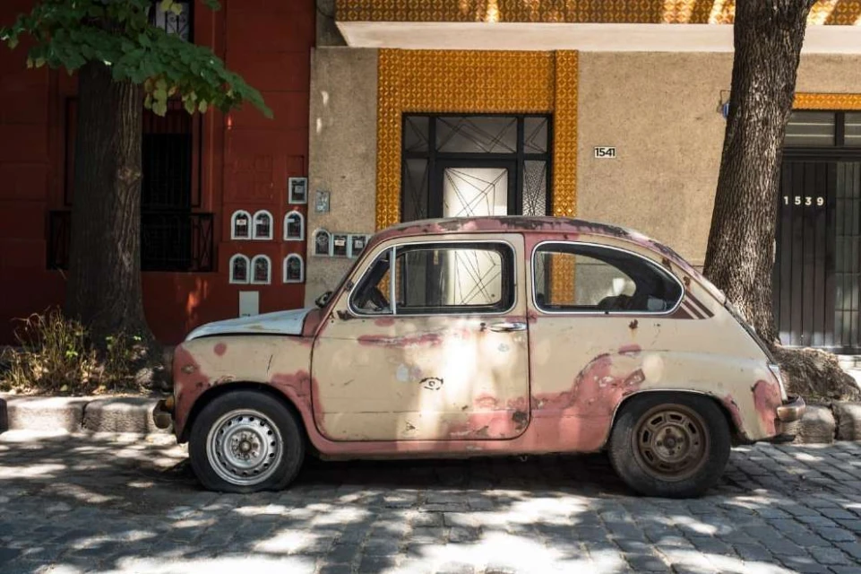 Vintage cream-colored car with flat tire parked on cobblestone street in front of orange and tan buildings with window details, trees nearby.