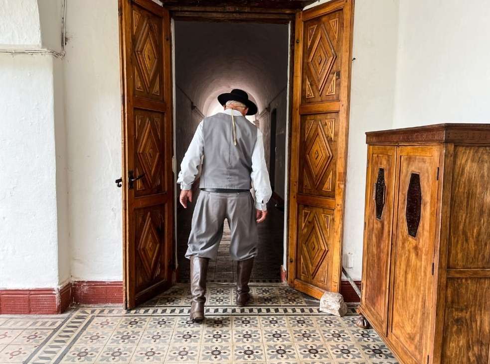 Man in gaucho attire stands in a doorway of an old building, wearing a black hat, vest, bombachas, and boots on a tiled floor.
