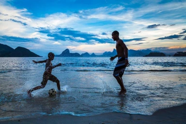 Two males playing soccer on a wet sandy beach at dusk with mountains and ocean in the background under a blue and white sky