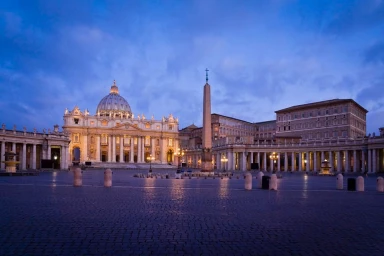 Wide evening view of St. Peter's Basilica and St. Peter's Square in Vatican City lit up under a blue twilight sky.