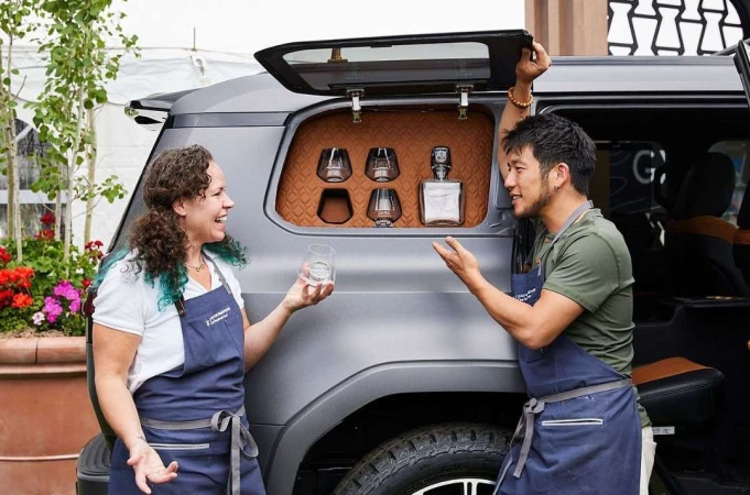 Two people stand by a gray SUV with an open hatch displaying glasses and a bottle, with a flower planter in the foreground.