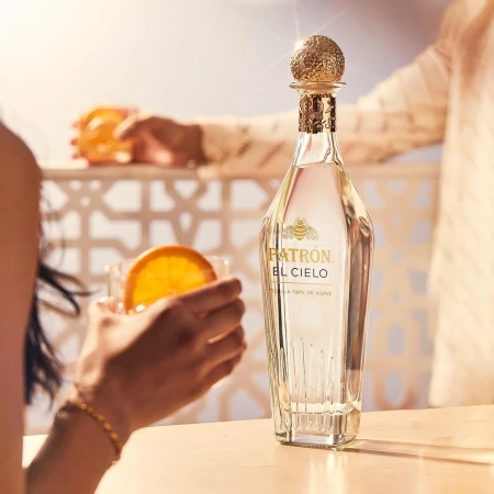 Bottle of Patron El Cielo tequila with a gold stopper on a counter with an orange slice and someone holding a drink in the background.