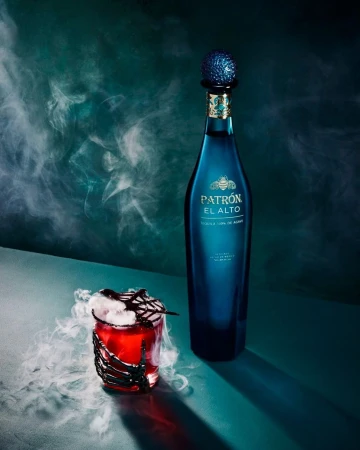 A bottle of Patron El Alto tequila and a cocktail glass with spooky Halloween decor on a green surface with a smoky background.