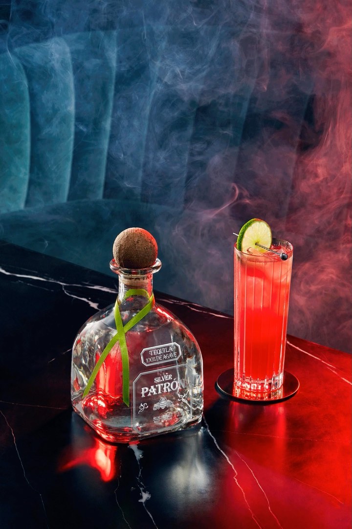 Patron Silver tequila bottle and cocktail glass with lime slice on a dark marble surface, illuminated by red and blue lighting with a smoky effect.