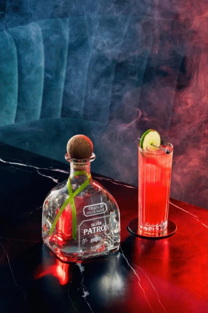 Patron Silver tequila bottle and cocktail glass with lime slice on a dark marble surface, illuminated by red and blue lighting with a smoky effect.