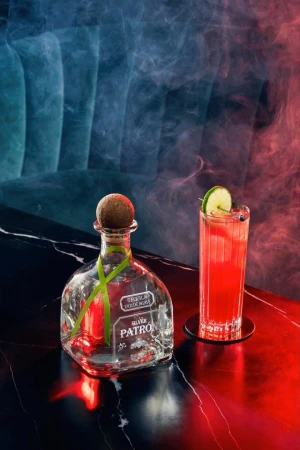 Patron Silver tequila bottle and cocktail glass with lime slice on a dark marble surface, illuminated by red and blue lighting with a smoky effect.