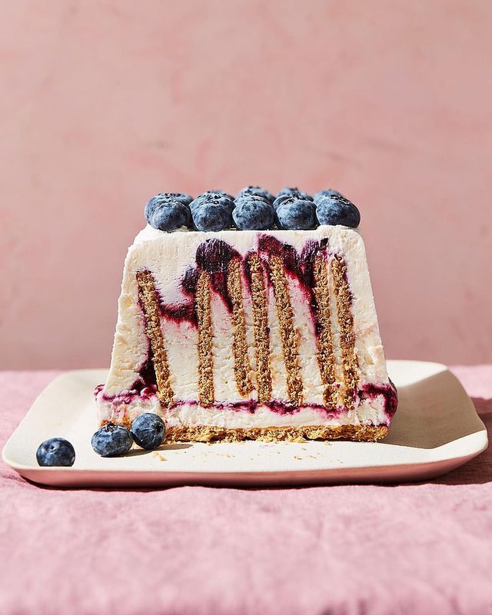 A slice of blueberry icebox cake with graham crackers and cream, sitting on a cream-colored plate against a light pink background.