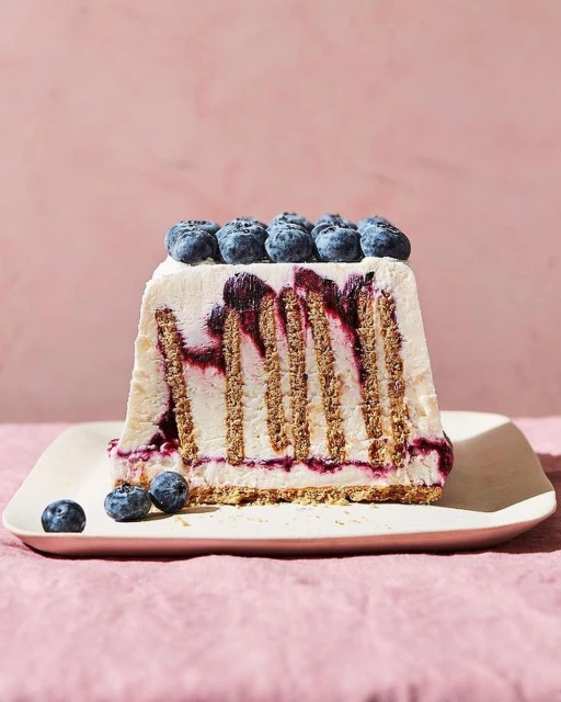 A slice of blueberry icebox cake with graham crackers and cream, sitting on a cream-colored plate against a light pink background.