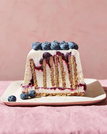 A slice of blueberry icebox cake with graham crackers and cream, sitting on a cream-colored plate against a light pink background.