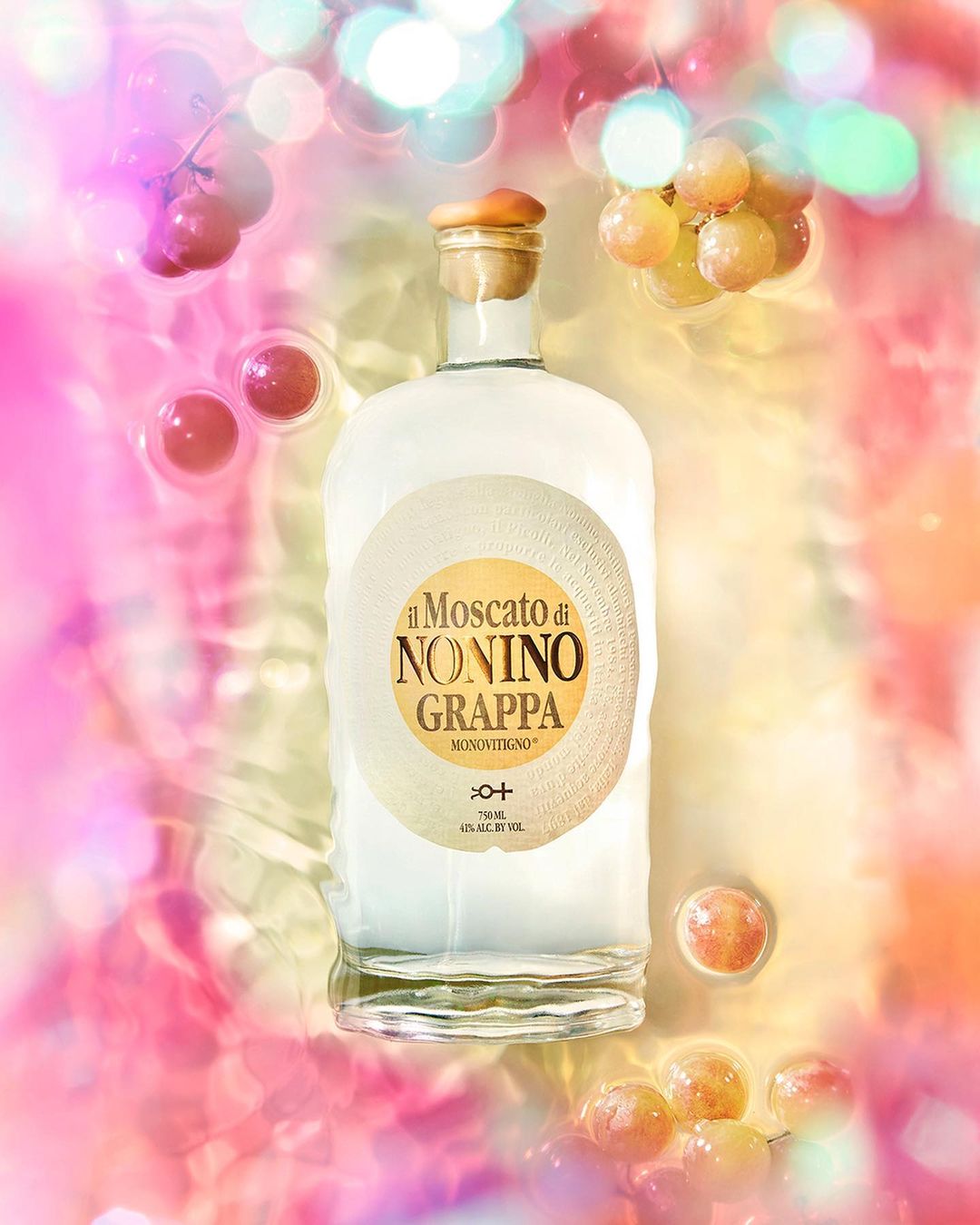 Bottle of Nonino Grappa with a cork stopper surrounded by floating grapes, set against a bokeh light and pink toned background.