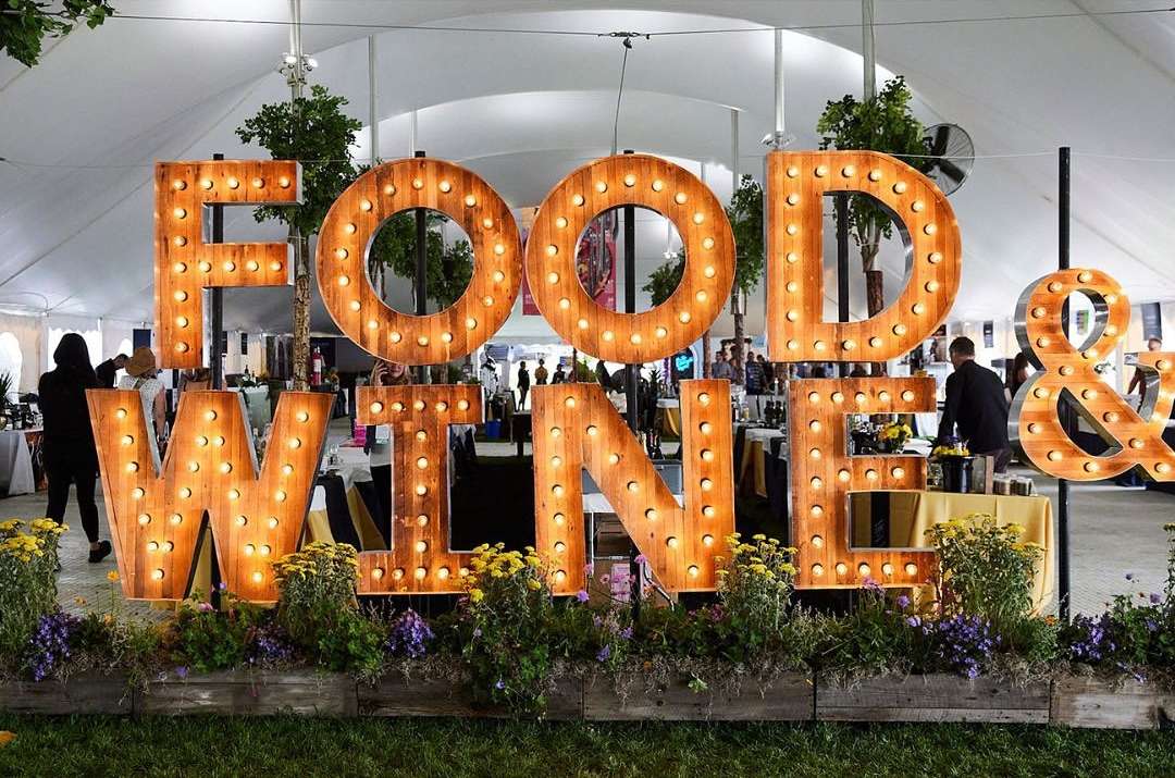Illuminated 'Food & Wine' sign at an outdoor event with flowers, tables, and blurred people in the background under a tent.