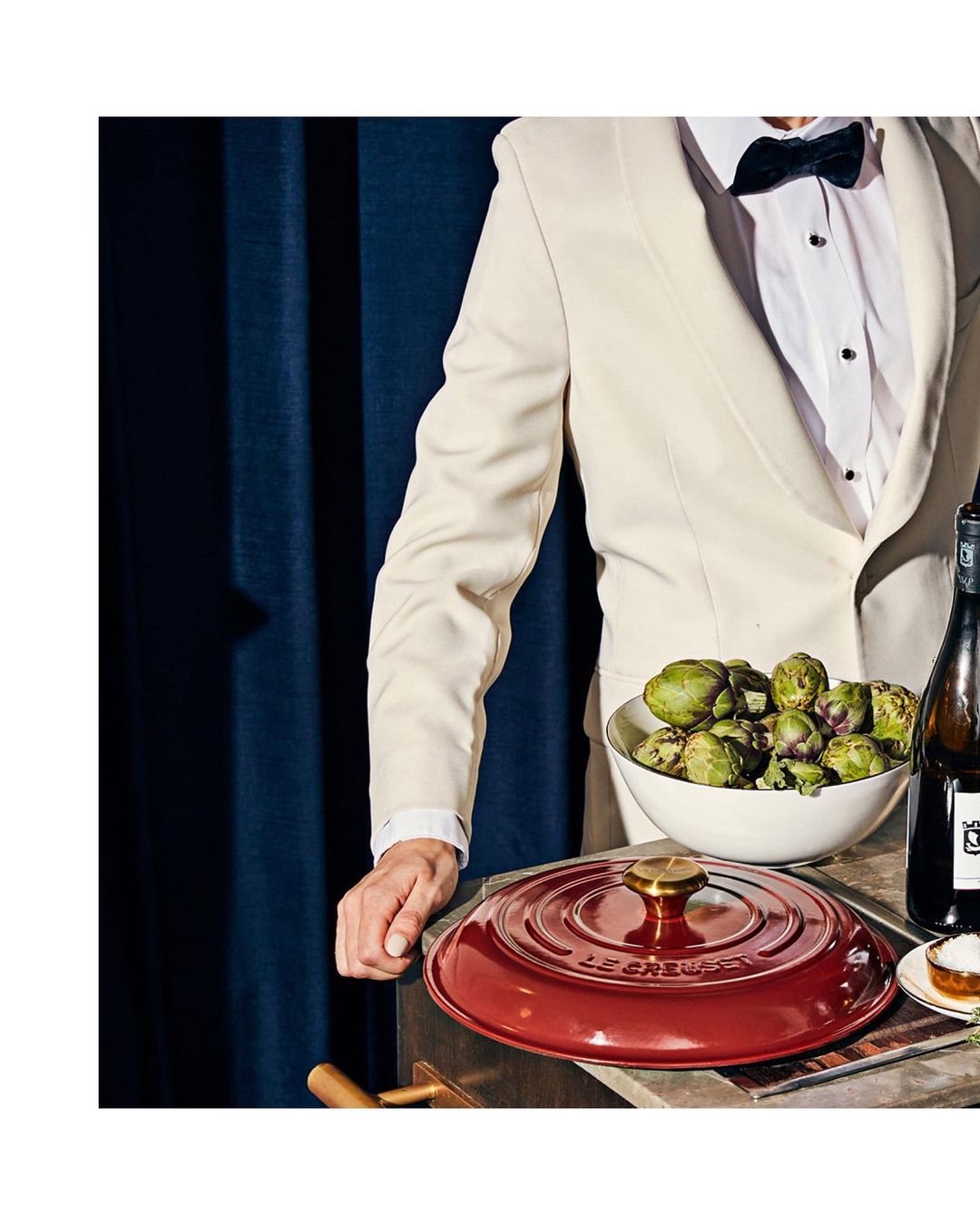 Person in a suit and bow tie standing next to a table with a red Le Creuset pot, artichokes in a bowl, and a bottle of wine.