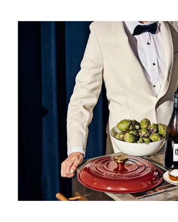 Person in a suit and bow tie standing next to a table with a red Le Creuset pot, artichokes in a bowl, and a bottle of wine.