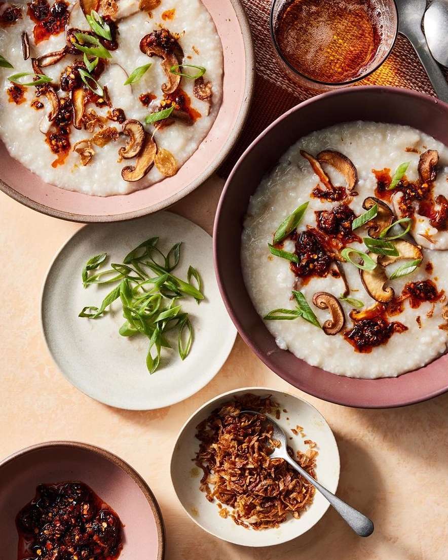 Overhead shot of two bowls of congee topped with mushrooms, chili oil, and scallions, accompanied by bowls of condiments and sliced scallions.