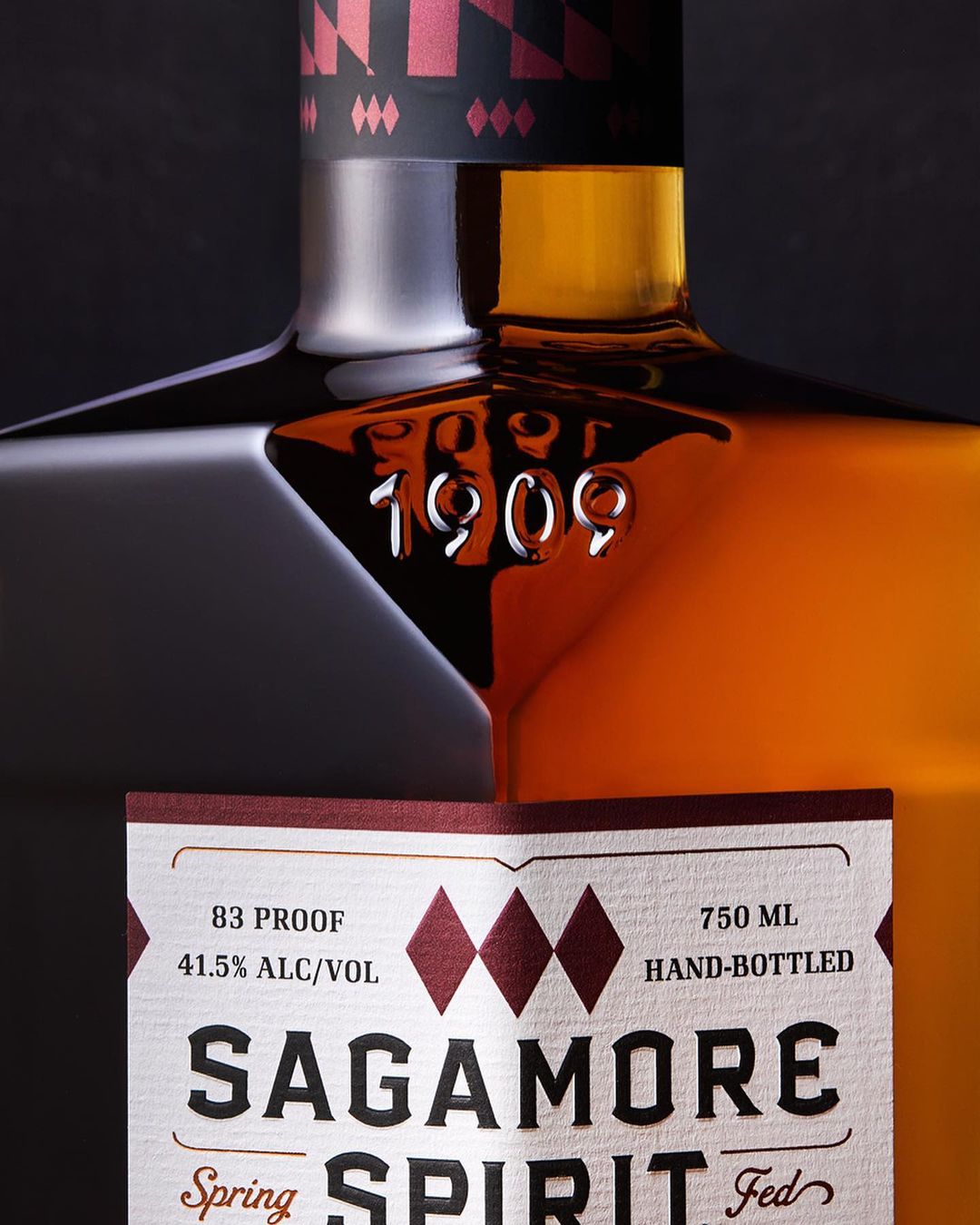 Close-up of a bottle of Sagamore Spirit, highlighting the brand name, 1909 embossing, and details on the label against a dark background.