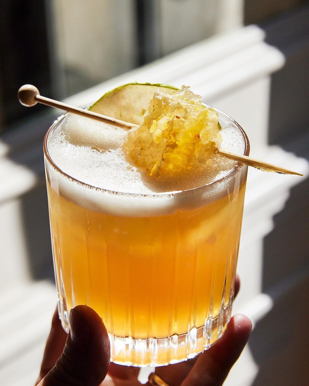 Cocktail drink with foamy top and honeycomb garnish held in hand against background with white molding and natural light.