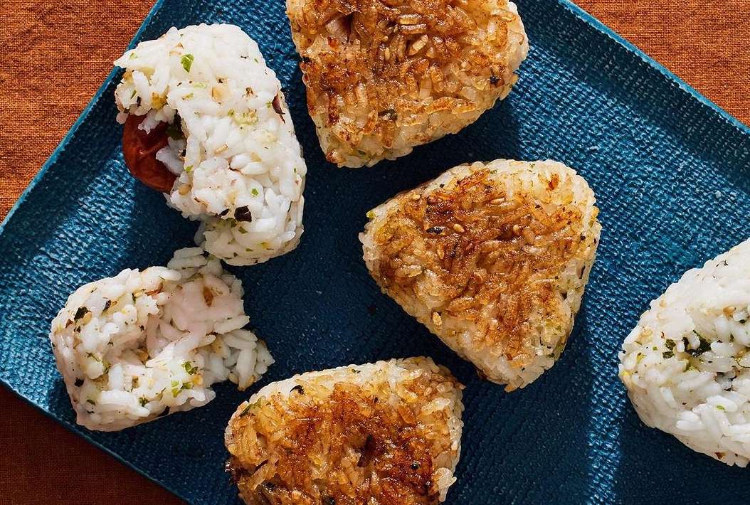 Overhead shot of six triangle-shaped onigiri (Japanese rice balls) on a blue textured surface, some seasoned and some plain.