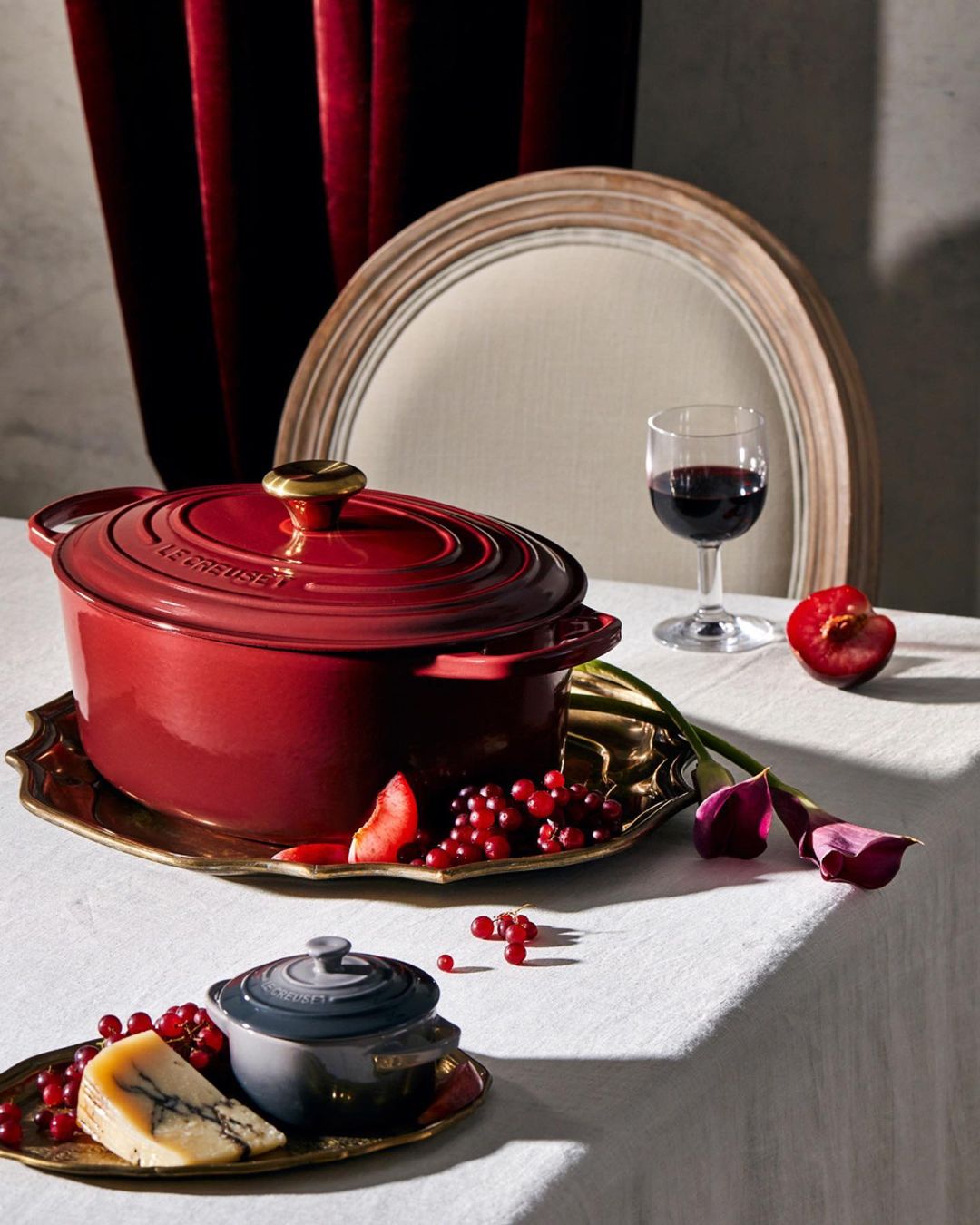 Red and gray Le Creuset pots on a table with a glass of red wine, cheese, red currants, a plum, tulips, and a chair in the background.