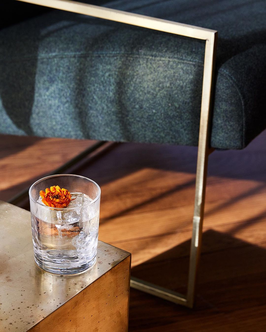 Cocktail with ice and a flower garnish on a golden side table, alongside an upholstered chair with brass frame legs indoors.