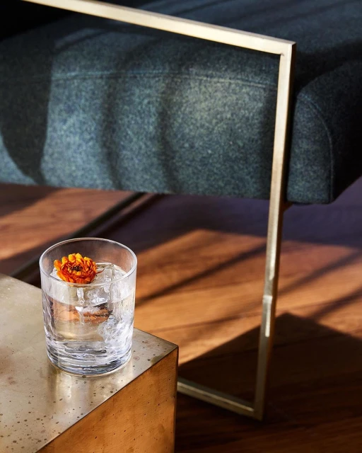 Cocktail with ice and a flower garnish on a golden side table, alongside an upholstered chair with brass frame legs indoors.