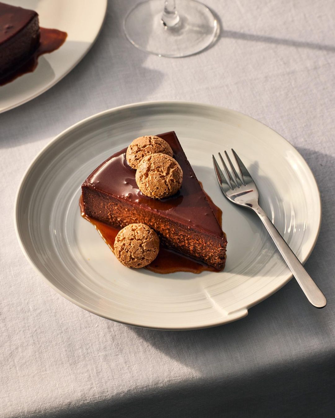 Slice of chocolate cake with glaze, topped with cookies on a white plate, alongside a fork and another slice in background.