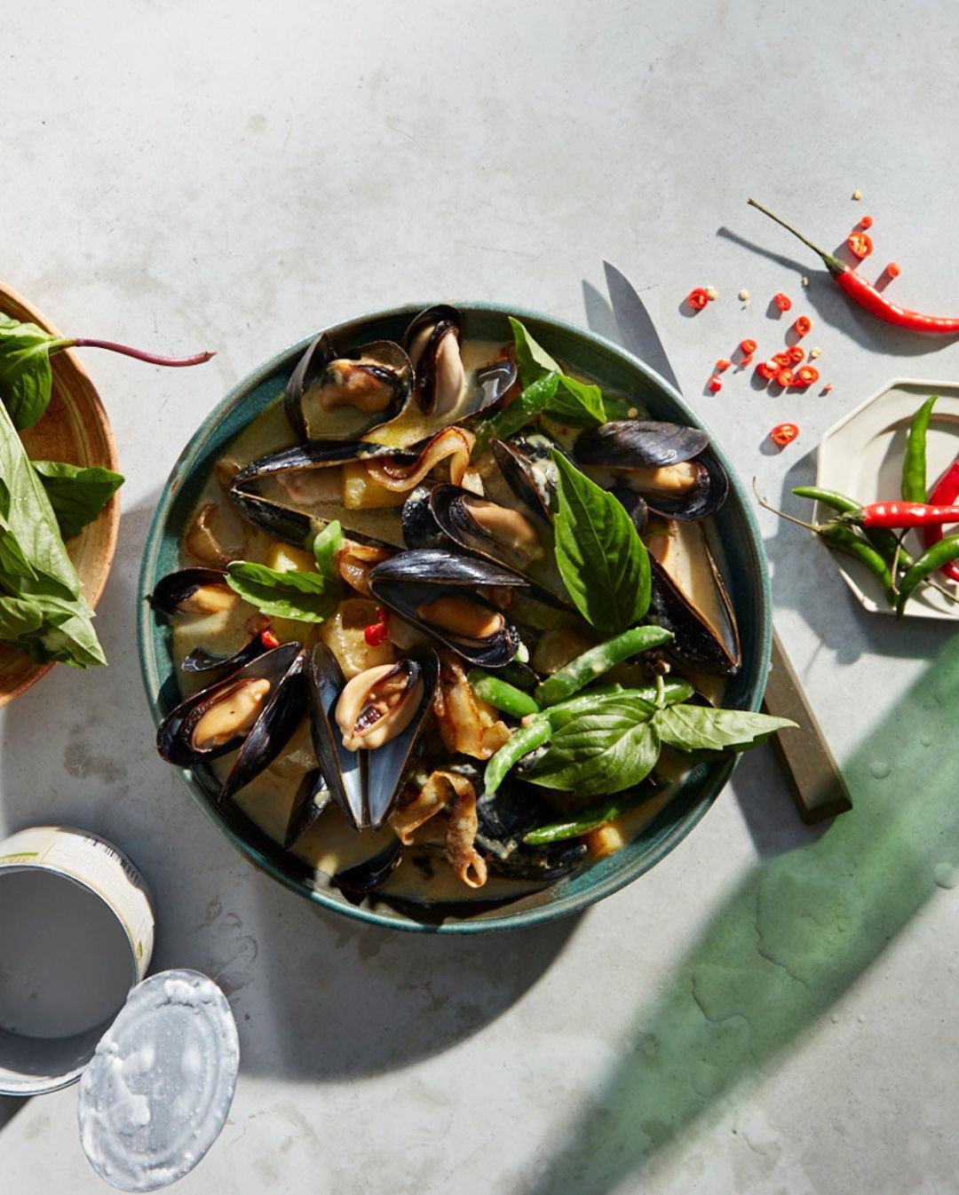 Overhead shot of mussel and vegetable stew with spices, served in teal bowl, with wooden bowl of leafy greens and red chilis.