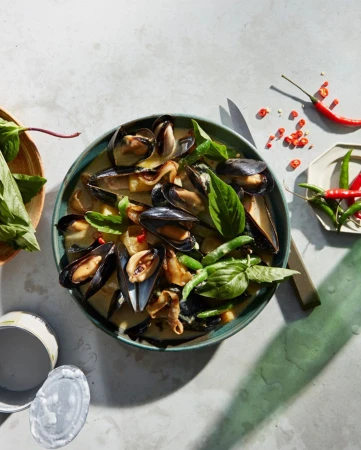 Overhead shot of mussel and vegetable stew with spices, served in teal bowl, with wooden bowl of leafy greens and red chilis.