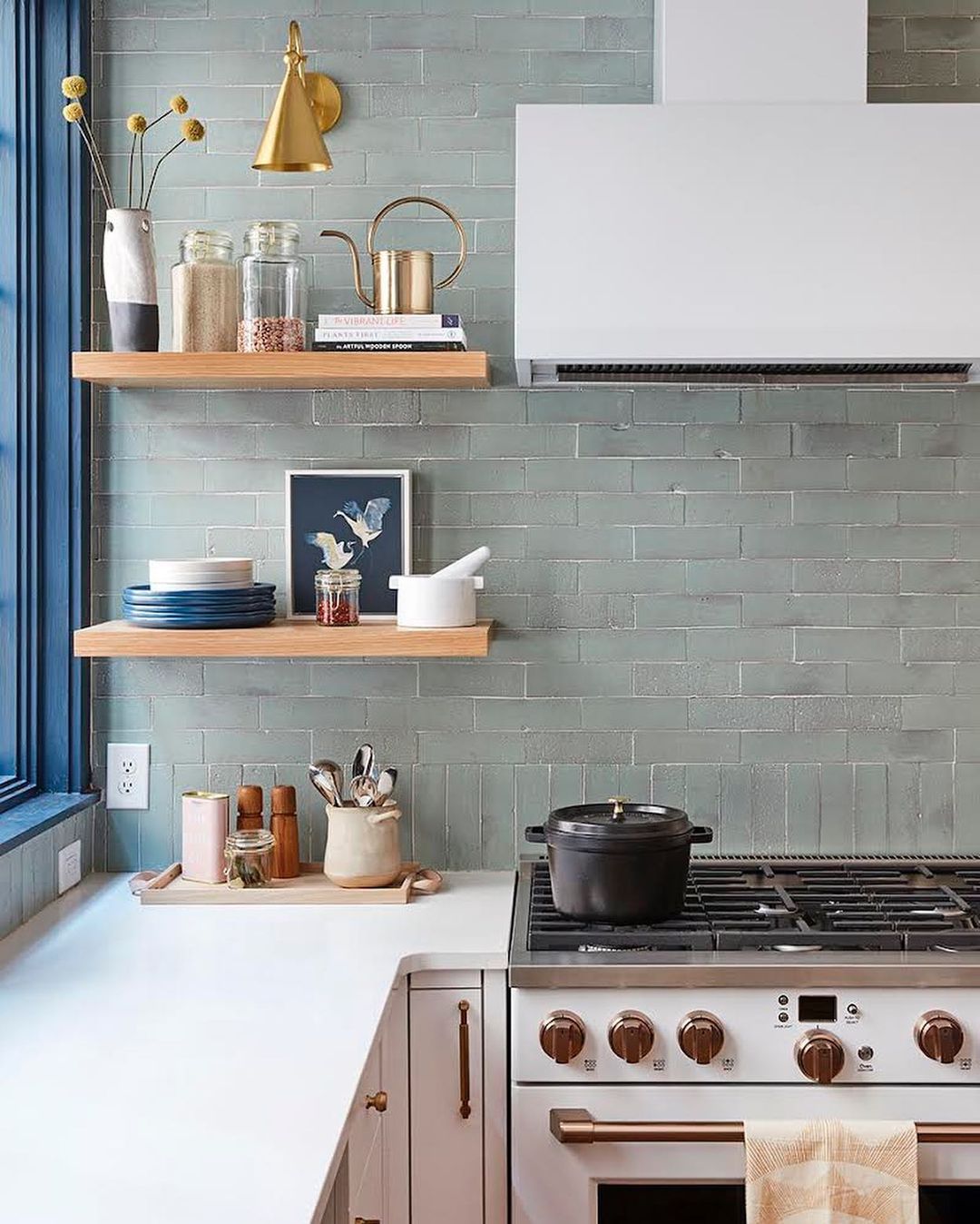 Interior shot of a modern kitchen featuring a stove with a pot, shelves with jars and ceramics, and a tile backsplash.