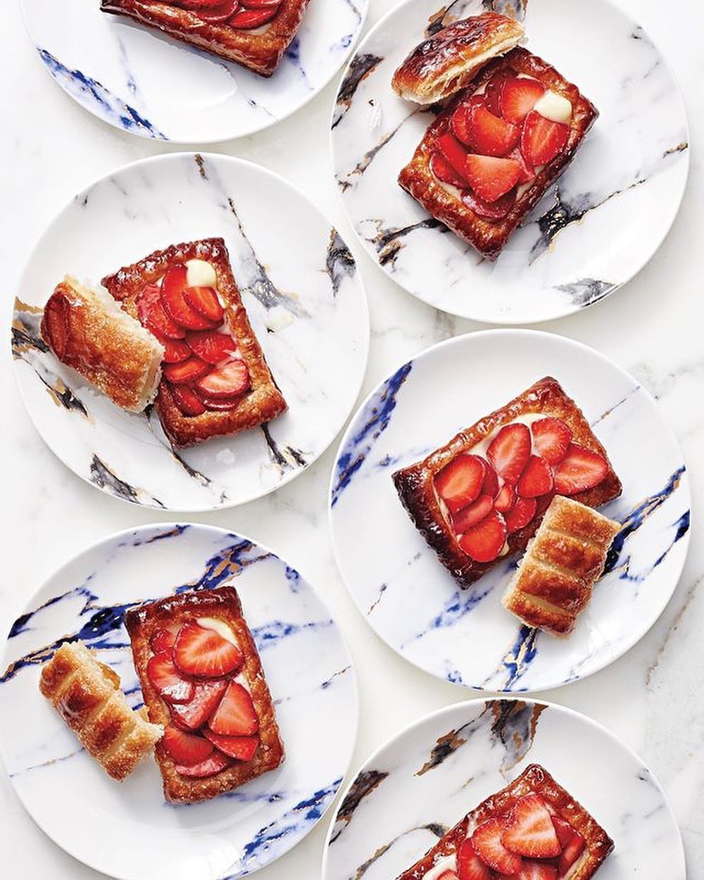 Overhead view of six marble plates, each with a rectangular strawberry tartlet and a pastry roll, showcasing food presentation.
