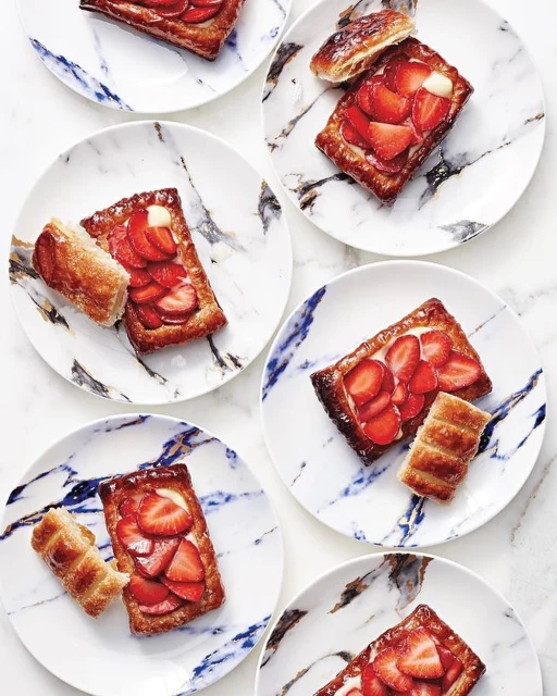 Overhead view of six marble plates, each with a rectangular strawberry tartlet and a pastry roll, showcasing food presentation.