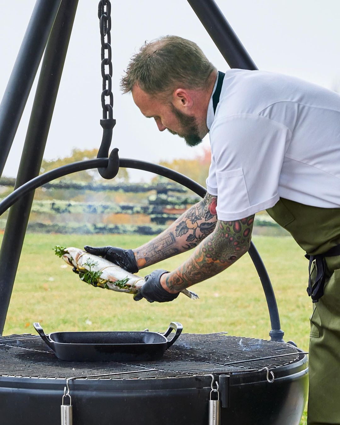 Chef in a white coat prepares a fish dish outside on a grill with an outdoor oven during the daytime in a food preparation scene.