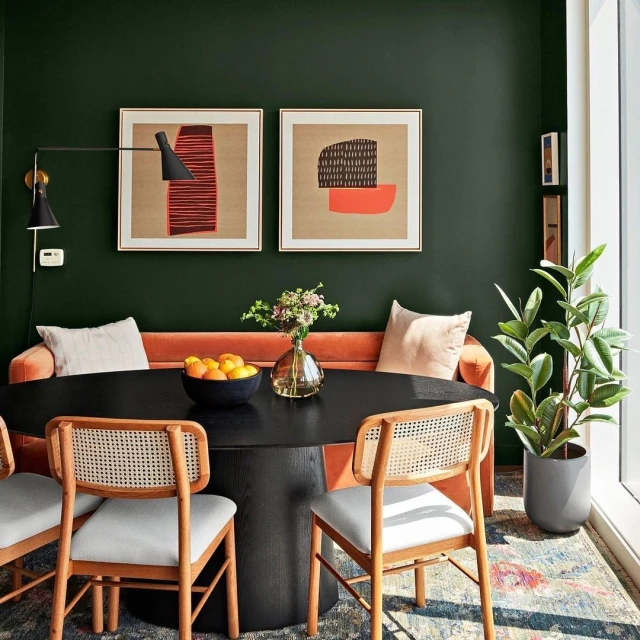 Interior design showcasing a modern dining area with a black table, woven chairs, an orange sofa, and abstract art against a dark green wall.