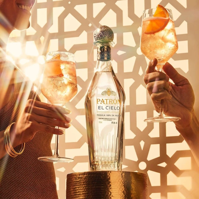Patrón El Cielo tequila bottle presented with two cocktails in stemmed glasses with orange slice garnish, against a patterned background.