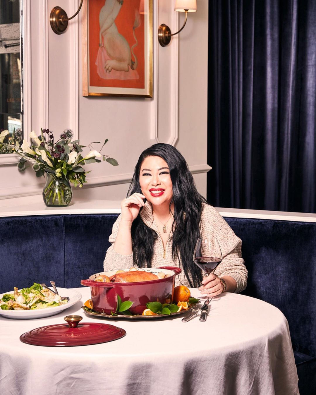 Smiling woman with long black hair sits at a round table in a restaurant, with a casserole dish, salad, wine, and flowers.