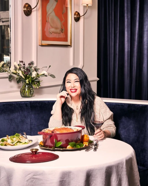 Smiling woman with long black hair sits at a round table in a restaurant, with a casserole dish, salad, wine, and flowers.