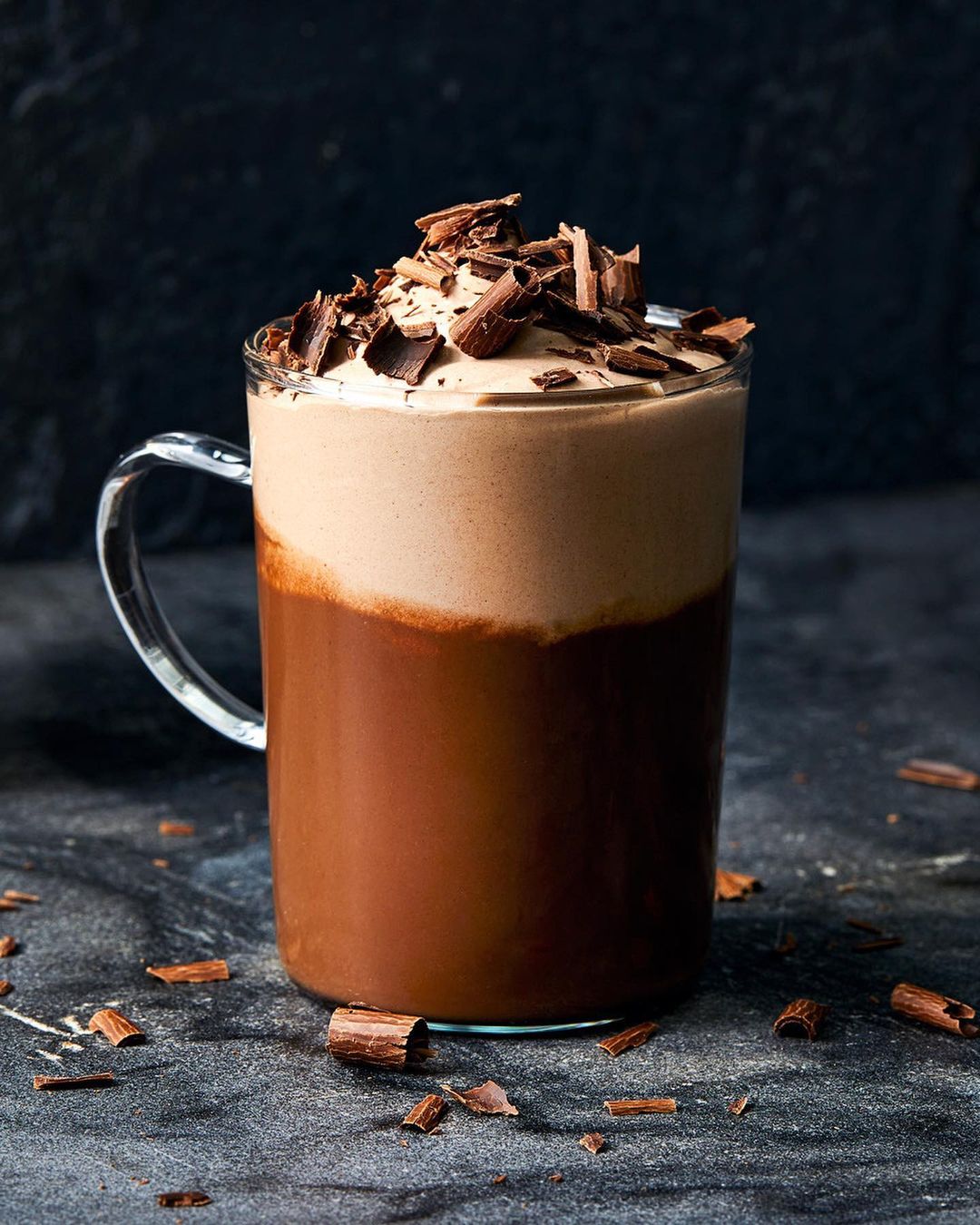 Close-up of a glass mug filled with a layered chocolate drink topped with whipped cream and chocolate shavings on a dark surface.