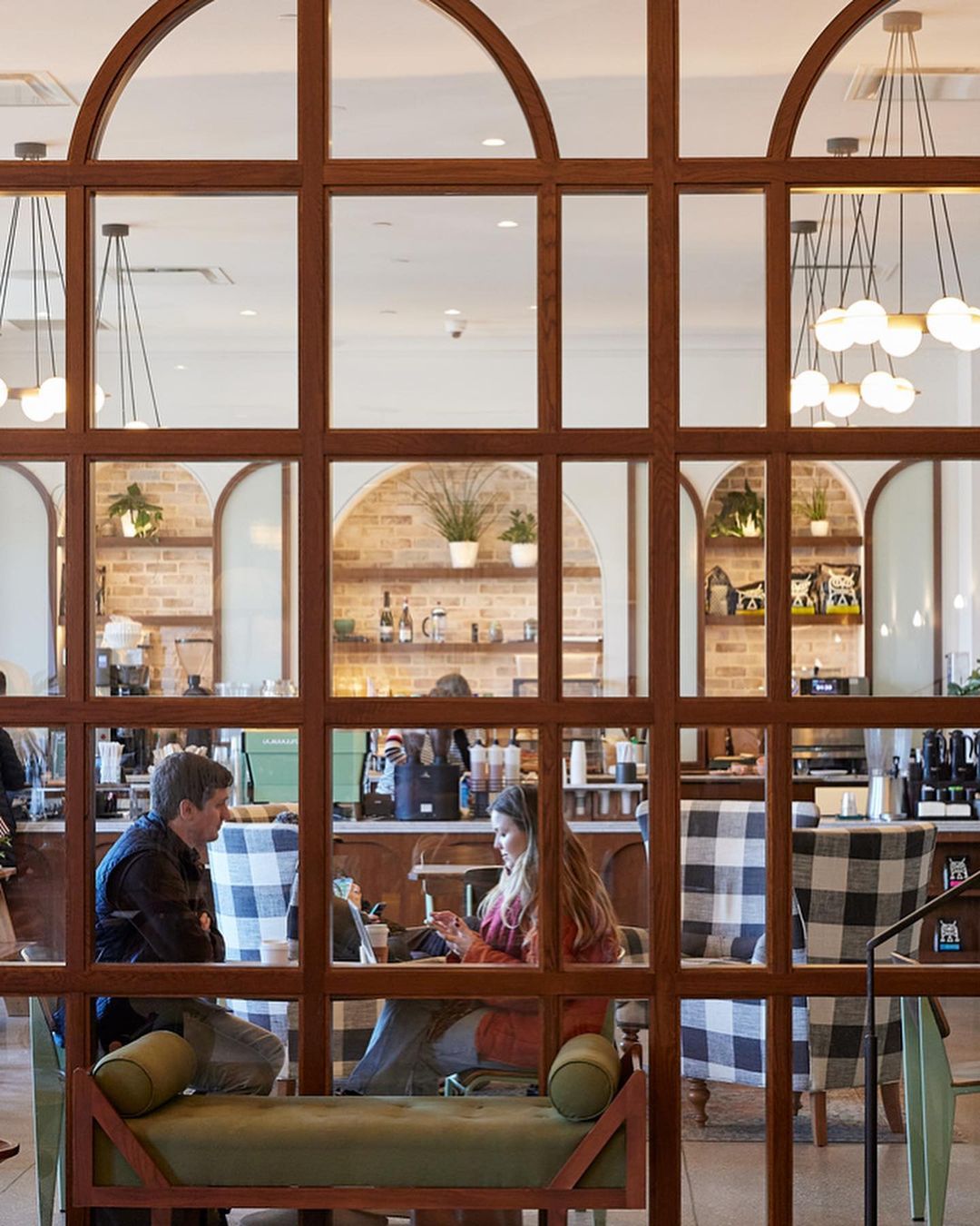 People sit at tables inside a bright cafe with modern lighting and decor, viewed through a decorative wood and glass window frame.