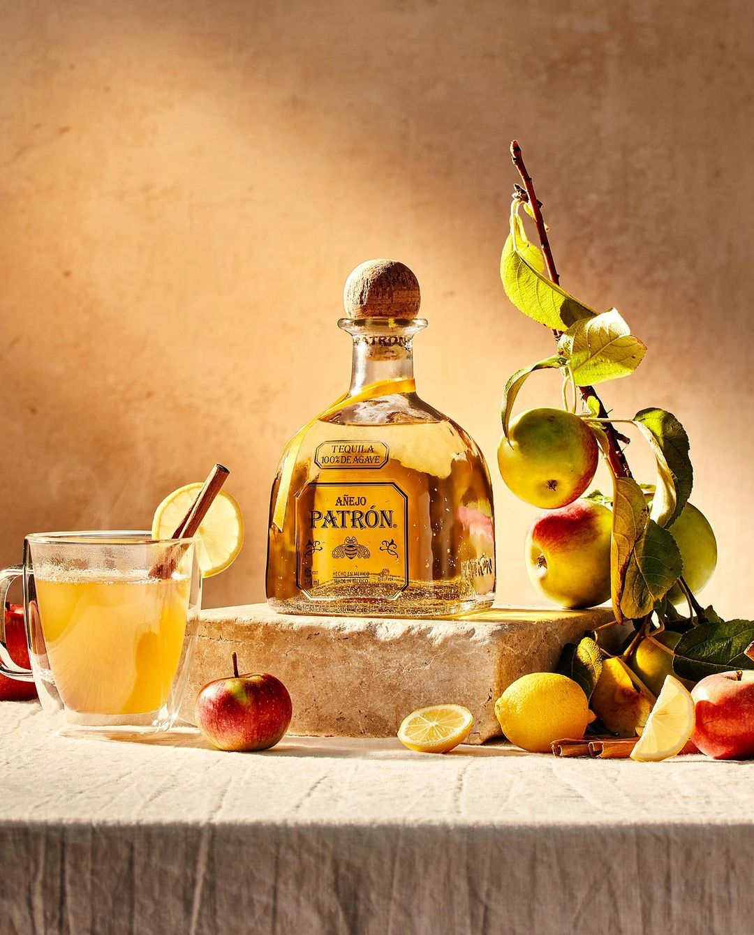 Still life featuring a Patron tequila bottle, glass of apple cider with cinnamon stick, apples, and lemons on a textured surface.