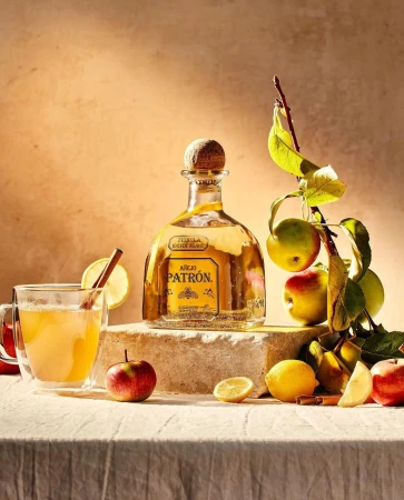 Still life featuring a Patron tequila bottle, glass of apple cider with cinnamon stick, apples, and lemons on a textured surface.