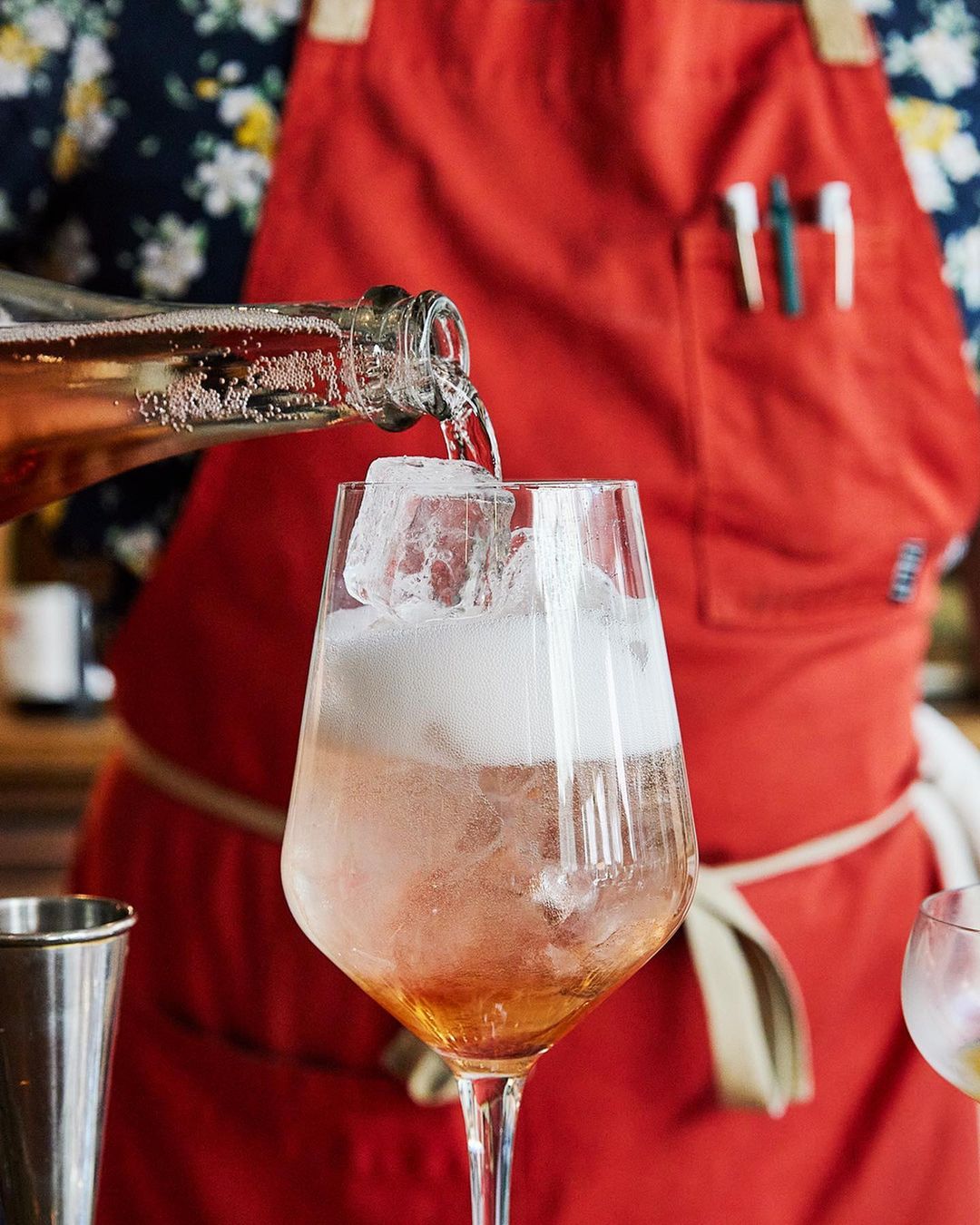 Bartender pours sparkling beverage from a clear bottle into an elegant glass filled with ice and a pink-tinted liquid.