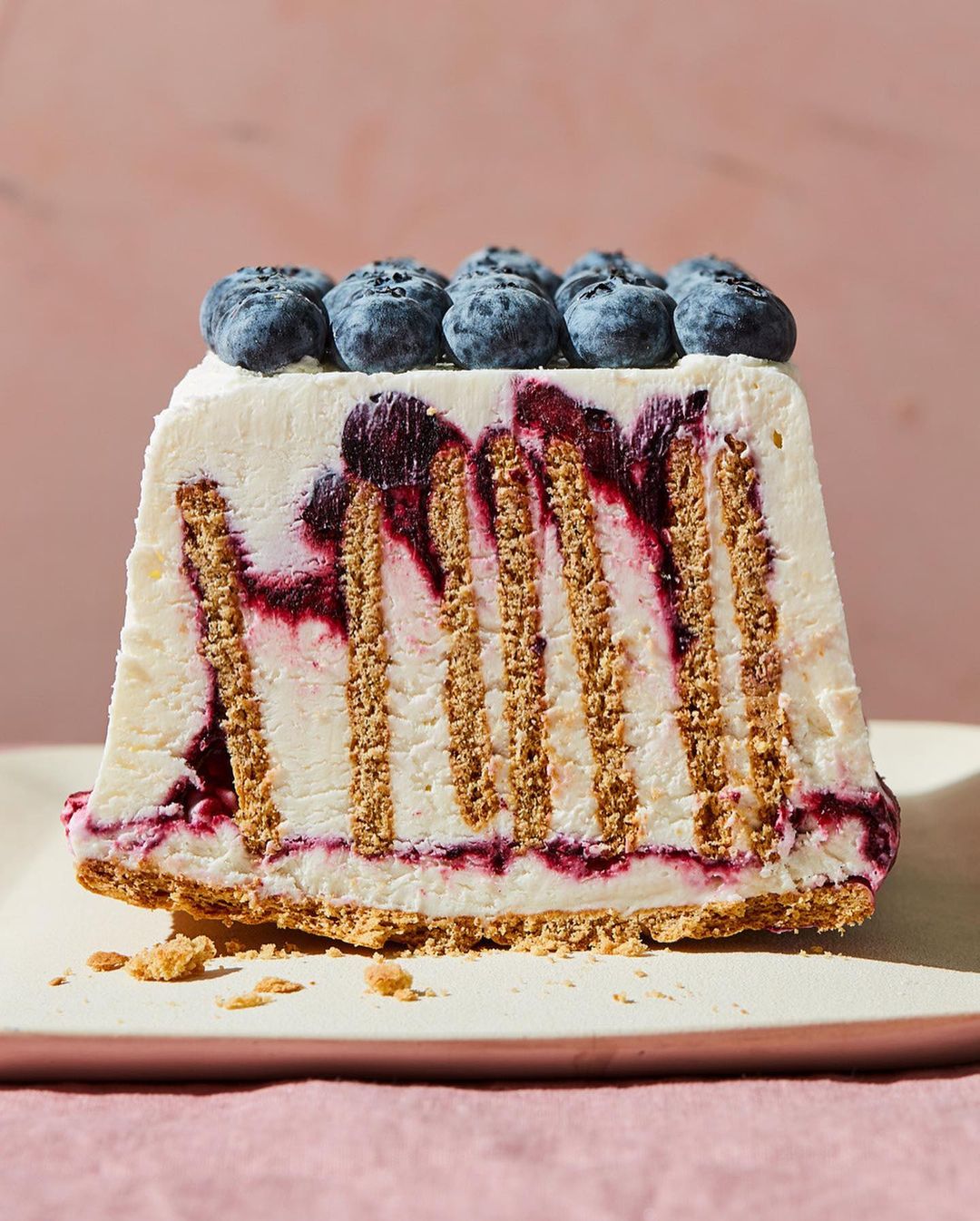 Close-up shot of a rectangular slice of blueberry icebox cake with graham cracker layers, topped with fresh blueberries on a pink background.