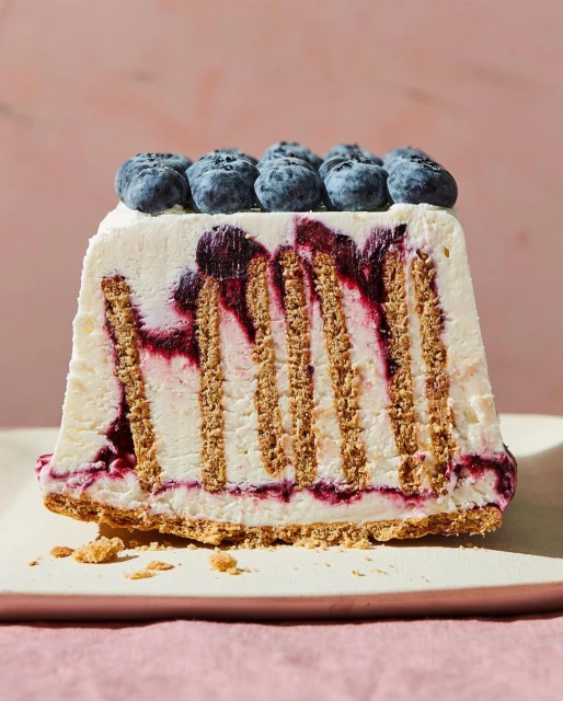 Close-up shot of a rectangular slice of blueberry icebox cake with graham cracker layers, topped with fresh blueberries on a pink background.