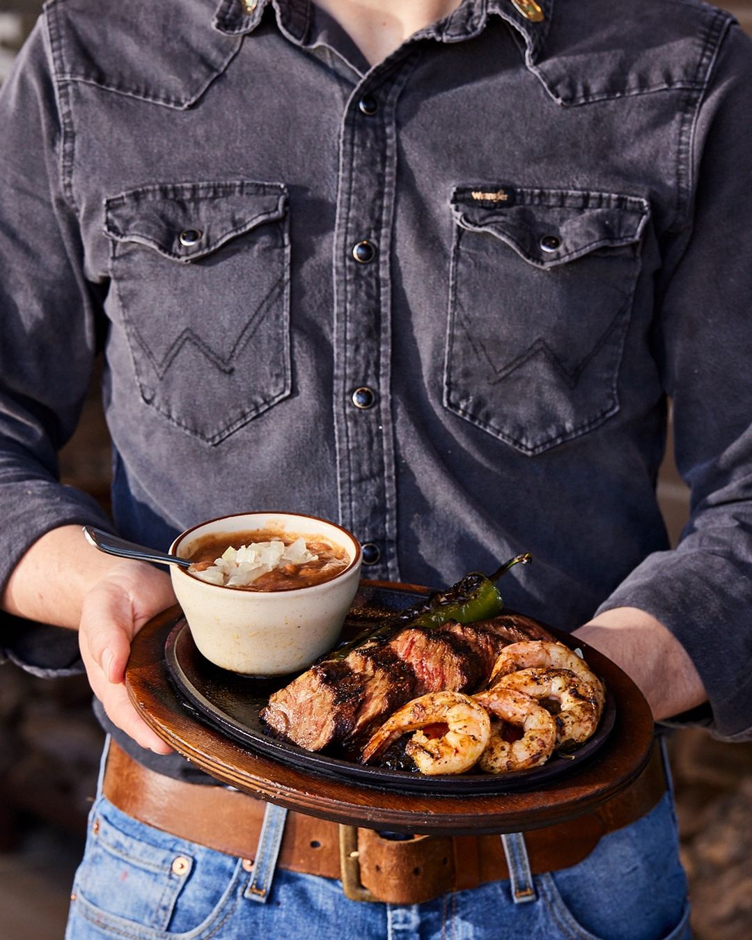 Person in denim shirt holds wooden platter of grilled shrimp, steak, pepper, and beans in a bowl, presented in medium shot.