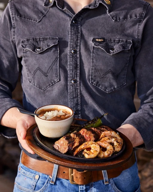 Person in denim shirt holds wooden platter of grilled shrimp, steak, pepper, and beans in a bowl, presented in medium shot.