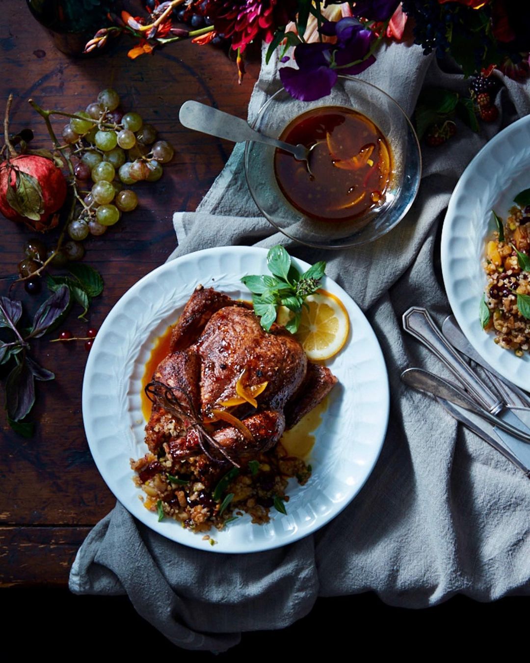 Overhead shot of a roast chicken dish with garnish on a white plate, next to a bowl of sauce, with flowers and fabric on a wooden table.