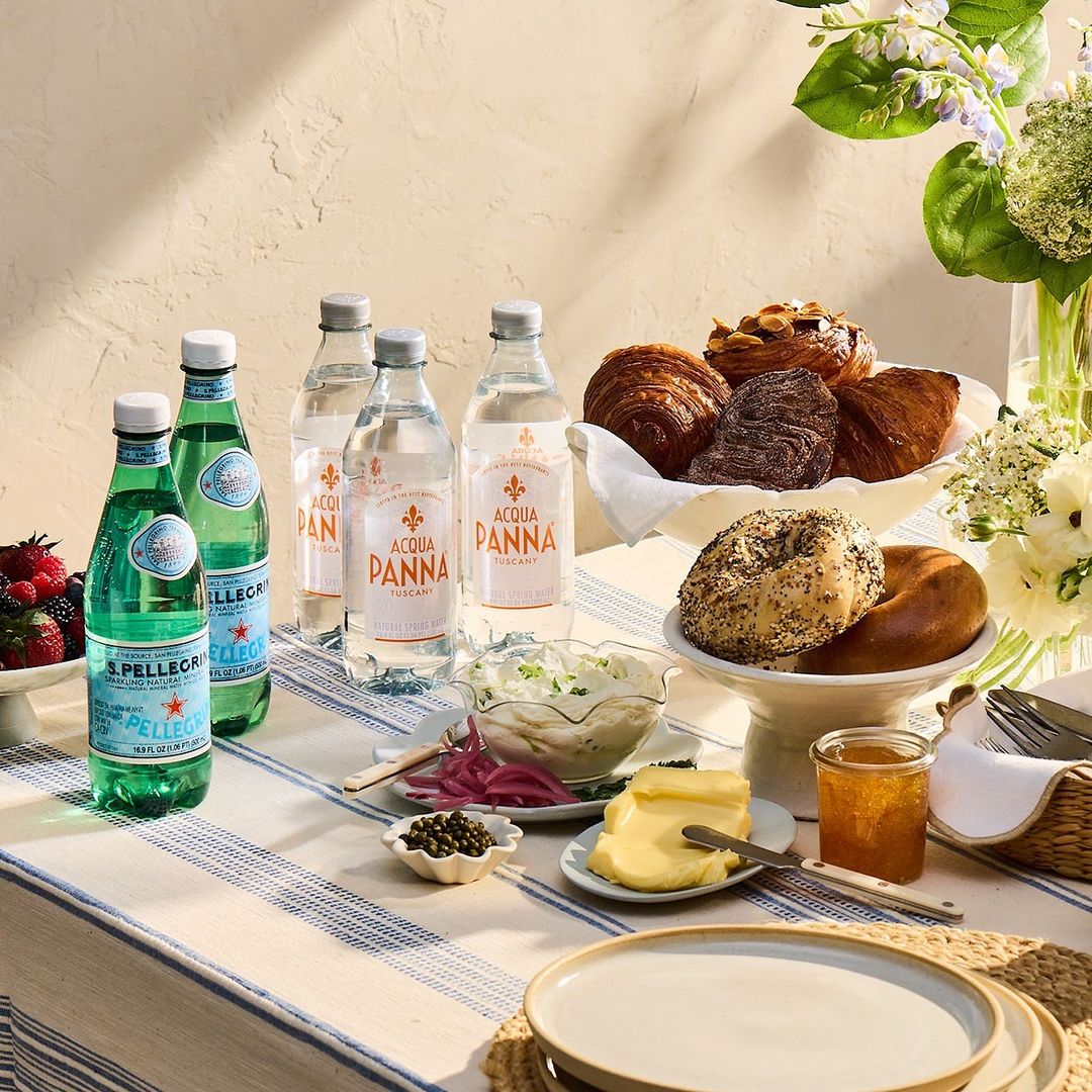 Outdoor picnic setup with bottled water, pastries, bagels, jam, butter, salad, flowers on a striped tablecloth under natural light.