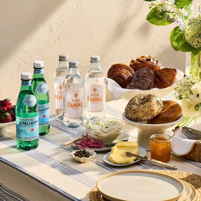 Outdoor picnic setup with bottled water, pastries, bagels, jam, butter, salad, flowers on a striped tablecloth under natural light.