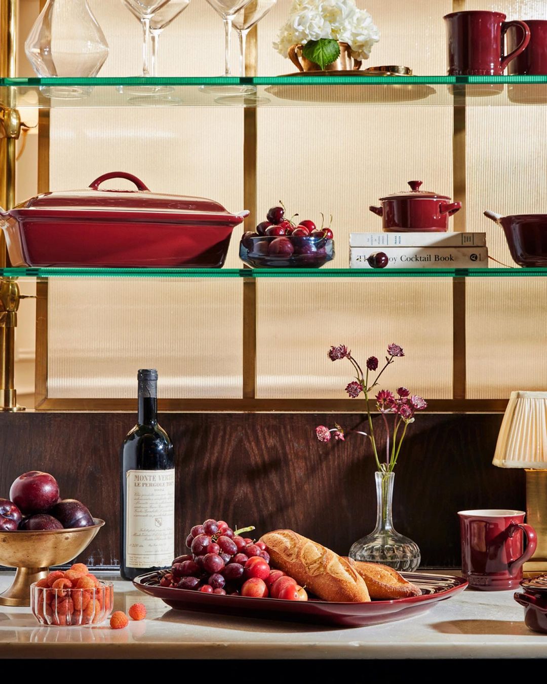 Still life composition featuring fruits, bread, wine, flowers and kitchenware arranged on shelves and a counter, in a warm indoor setting.