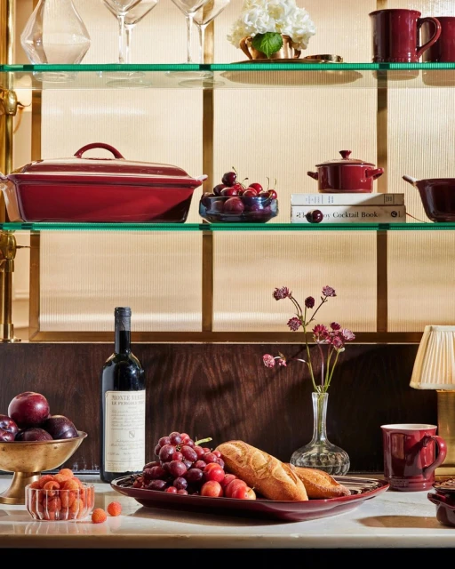 Still life composition featuring fruits, bread, wine, flowers and kitchenware arranged on shelves and a counter, in a warm indoor setting.